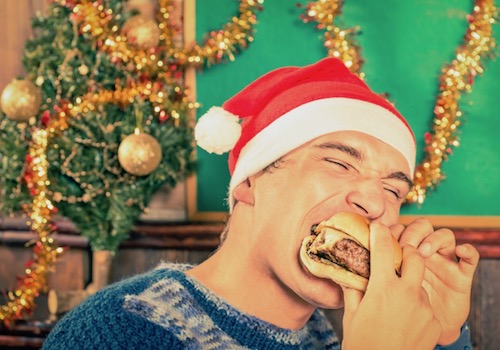 A Guy Wearing a Santa Hat Eating a Big Sandwich in Front of Xmas Decorations